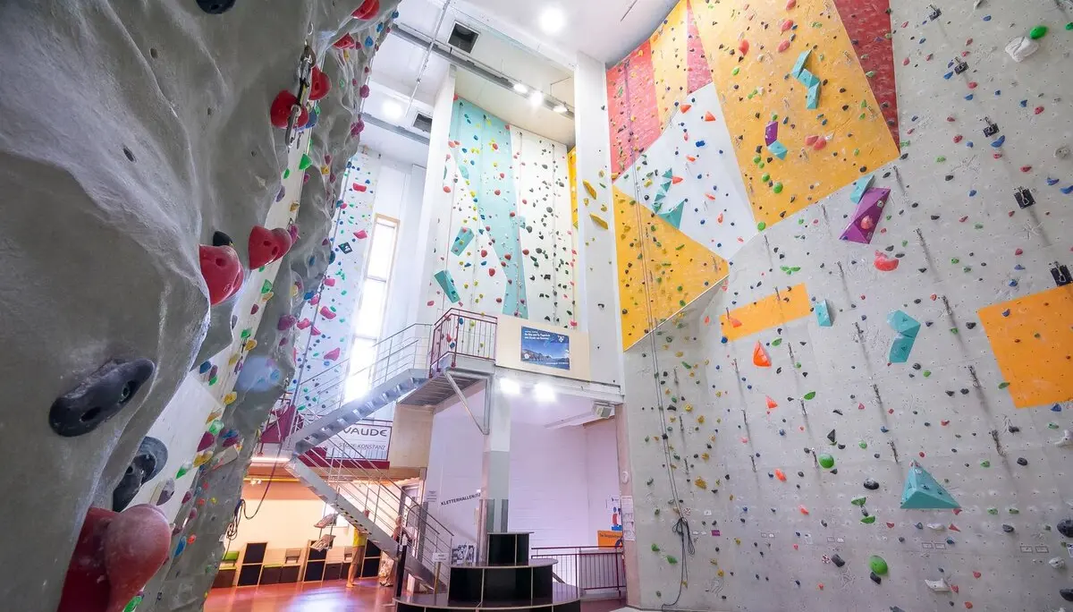 Innenansicht einer hellen Kletterhalle mit hohen, bunten Kletterwänden und überhängenden Elementen, Treppe in der Mitte, Sitzbereich unten. | © Test test