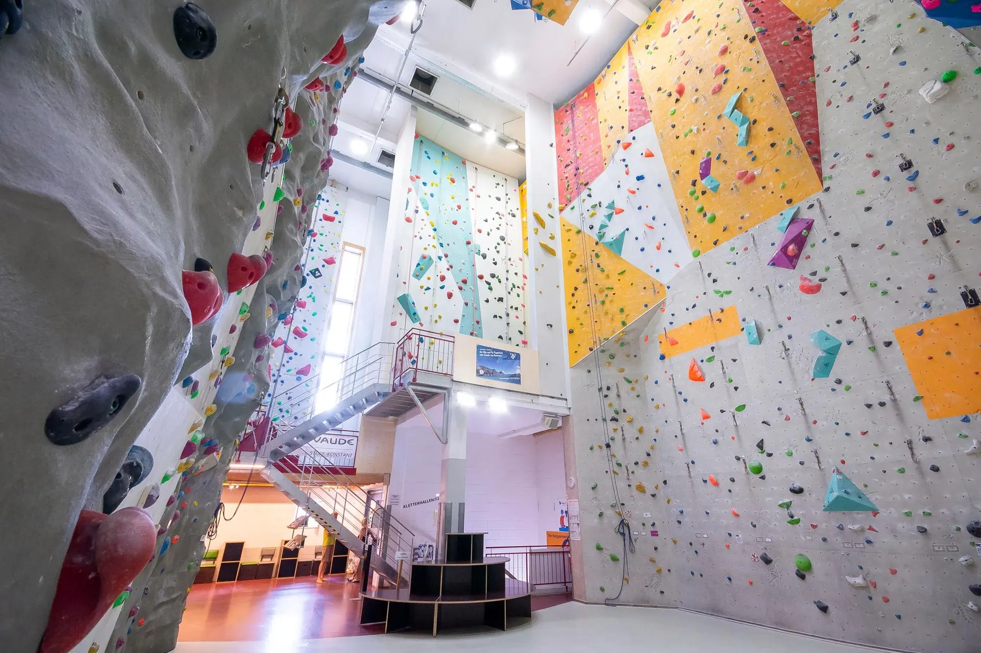 Innenansicht einer hellen Kletterhalle mit hohen, bunten Kletterwänden und überhängenden Elementen, Treppe in der Mitte, Sitzbereich unten. | © Test test