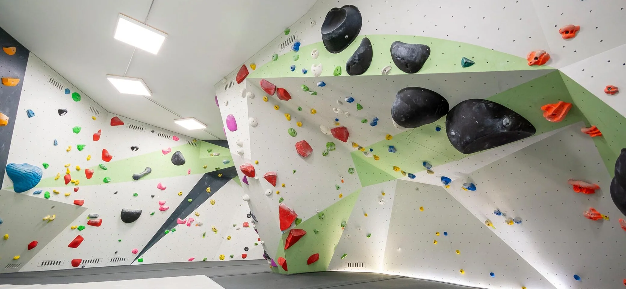 Helle Indoor-Boulderhalle mit geometrisch gestalteten Wänden, vielen bunten Klettergriffen und großen schwarzen Volumen, weicher Boden mit Matten. | © DAV Konstanz, Michael Dörfer