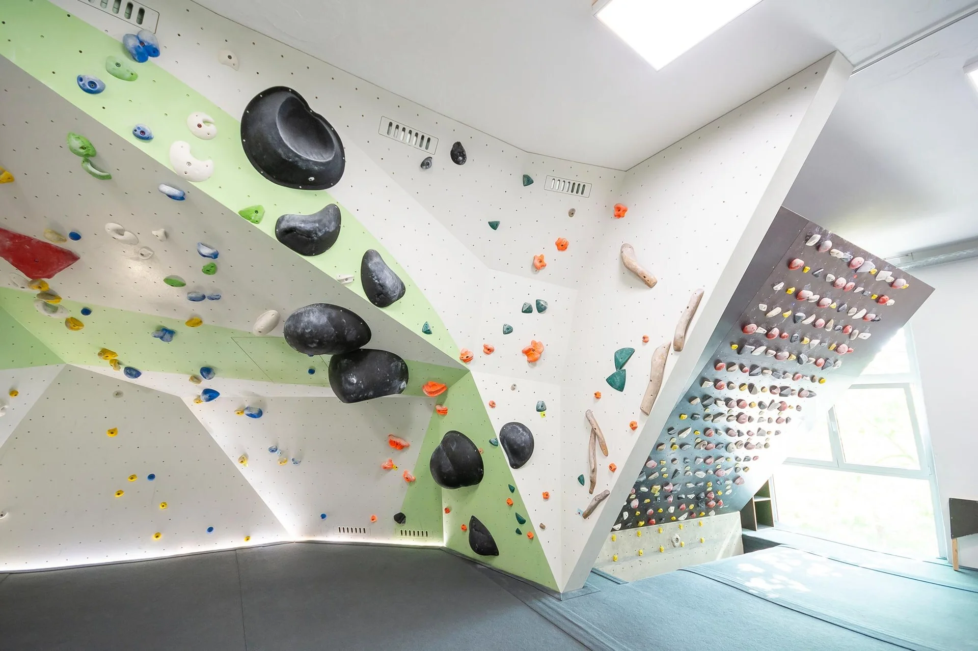 Helle Indoor-Boulderhalle mit geometrisch gestalteten Wänden, vielen bunten Klettergriffen und großen schwarzen Volumen, weicher Boden mit Matten. | © DAV Konstanz, Michael Dörfer