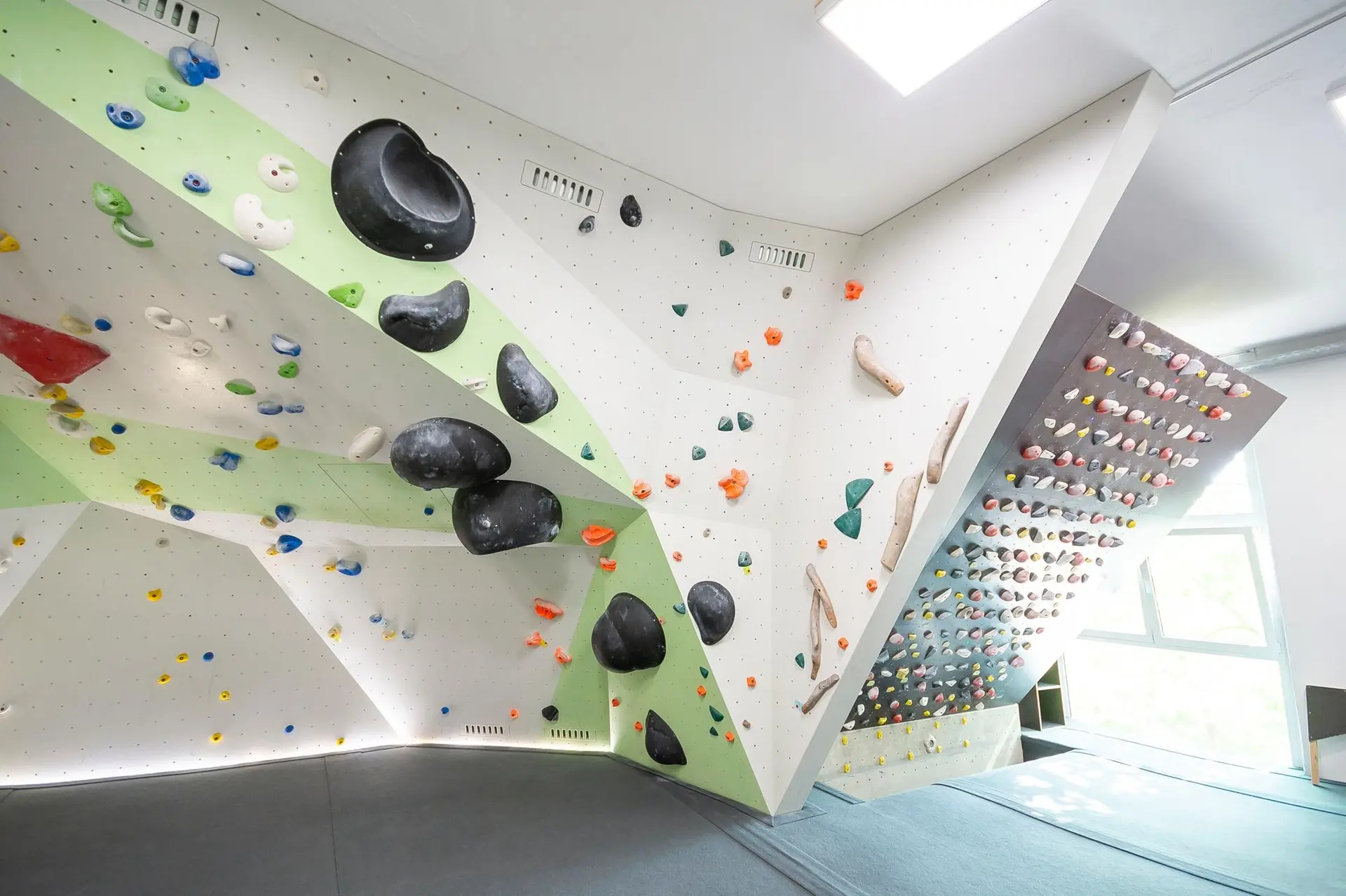 Helle Indoor-Boulderhalle mit geometrisch gestalteten Wänden, vielen bunten Klettergriffen und großen schwarzen Volumen, weicher Boden mit Matten. | © DAV Konstanz, Michael Dörfer