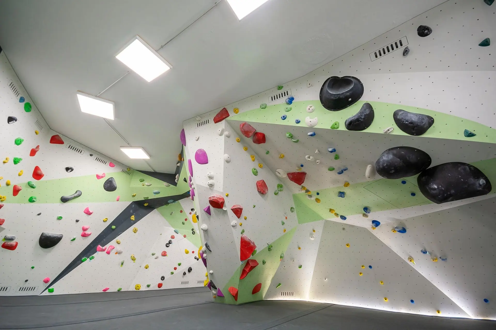 Helle Indoor-Boulderhalle mit geometrisch gestalteten Wänden, vielen bunten Klettergriffen und großen schwarzen Volumen, weicher Boden mit Matten. | © DAV Konstanz, Michael Dörfer